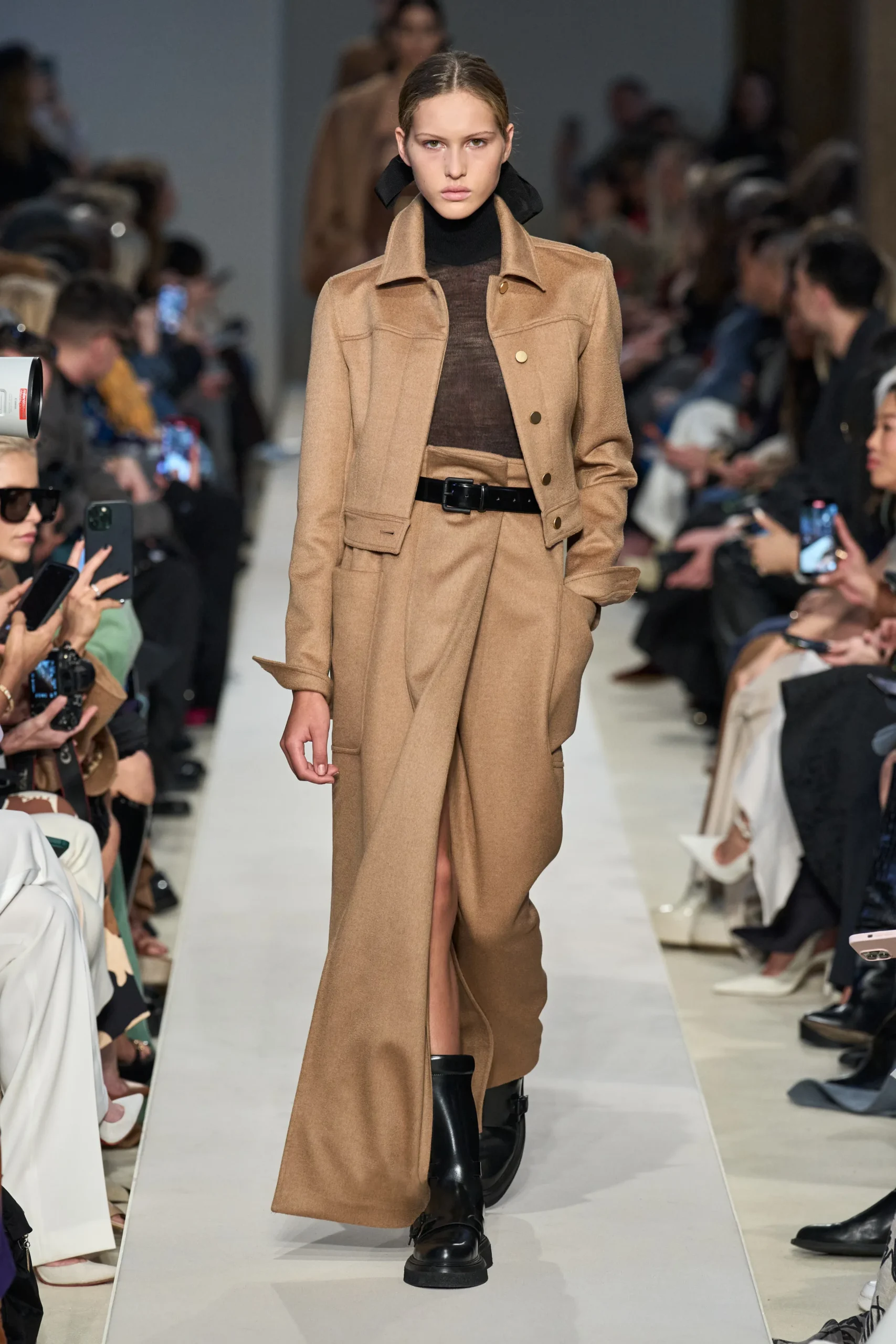 Model walking on a fashion runway, showcasing a fall ready-to-wear outfit by Max Mara. The ensemble features a tailored camel-coloured wool coat with large lapels and a matching wide-legged trouser, cinched at the waist with a black leather belt. Underneath, the model wears a dark turtleneck. She completes her look with black leather ankle boots. The audience, with their cameras out, lines the catwalk, capturing the moment.