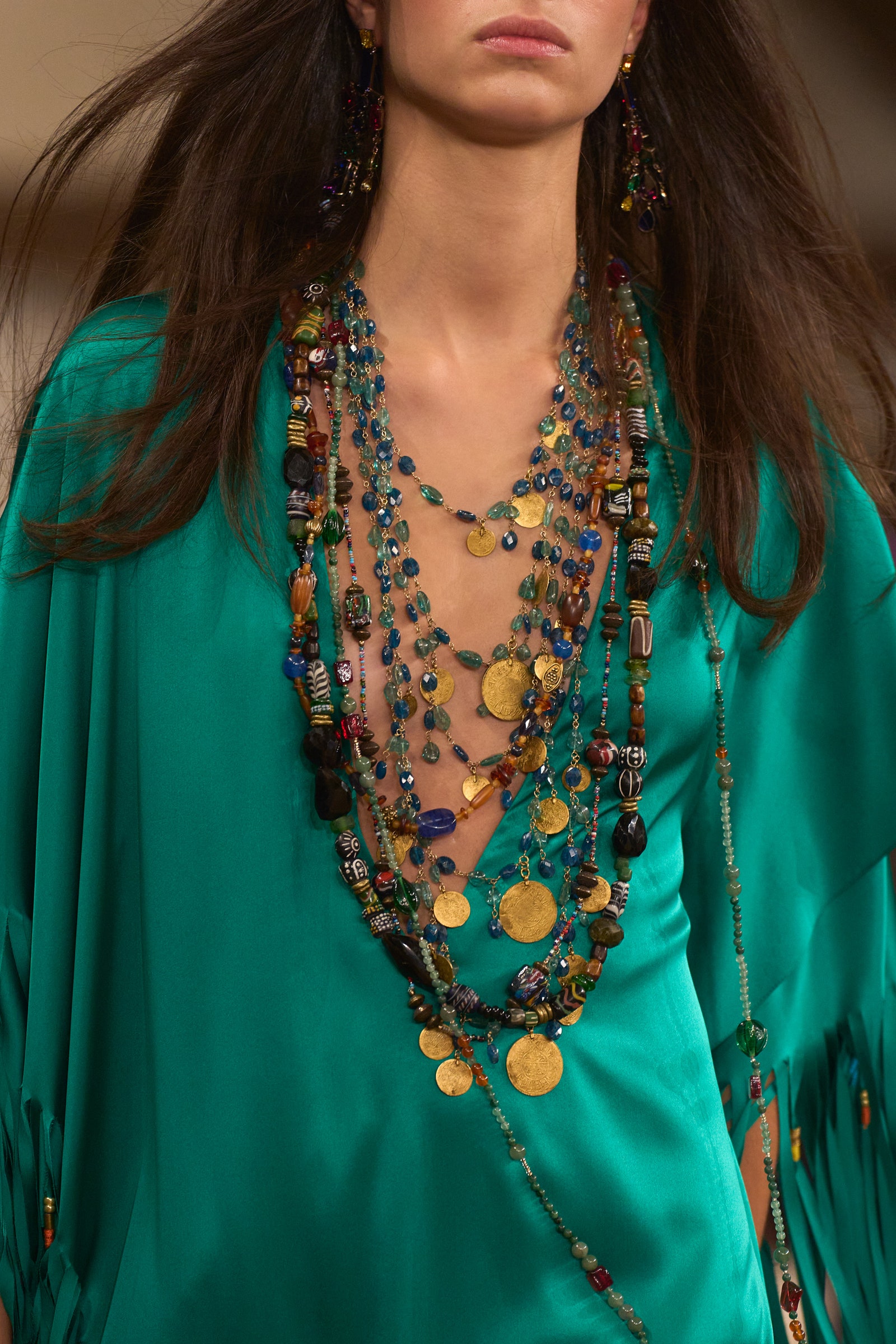 A cascade of vibrant, eclectic beads and coins flows in a symphony of colour against a striking teal backdrop. This detail from Ralph Lauren's Spring 2024 Ready-to-Wear collection captures a bohemian rhapsody, with layers of necklaces offering a rich palette of textures and hues, evoking the spirit of adventure and the art of personal expression.