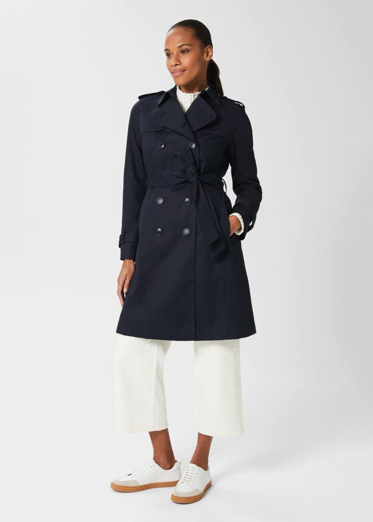 A model presents the Hobbs Saskia Shower Resistant Trench Coat in a classic navy hue. The coat's crisp lines and double-breasted design with a tie belt offer a flattering fit, while its timeless style is complemented by cropped white trousers and casual white trainers, epitomising transitional dressing with elegance and ease.