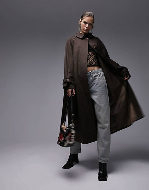 An image of a model striking a dynamic pose in a Topshop long-lined belted brushed trench coat in a rich chocolate hue. The coat is styled with an edgy lace top, relaxed grey trousers, and sleek black ankle boots. The look is completed with a graphic tote bag, showcasing a fashion-forward and layered autumn ensemble.