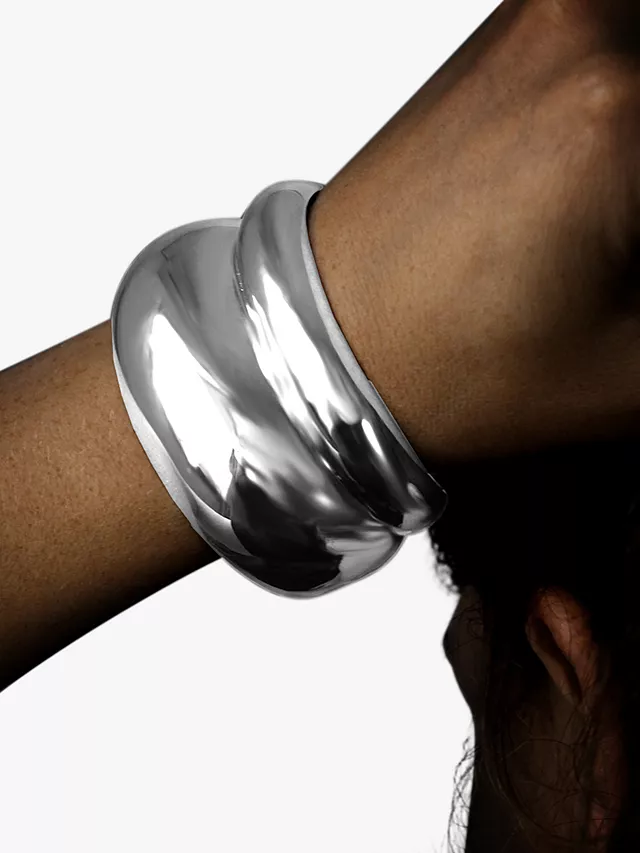 A bold and sculptural Nina B Polished Cumulus Cuff Bangle in gleaming silver, gracefully wraps around the wrist, reflecting light and exuding modern sophistication. The piece, with its fluid, organic shape, is a standout accessory available at John Lewis, combining both artistry and a touch of luxury within its design.