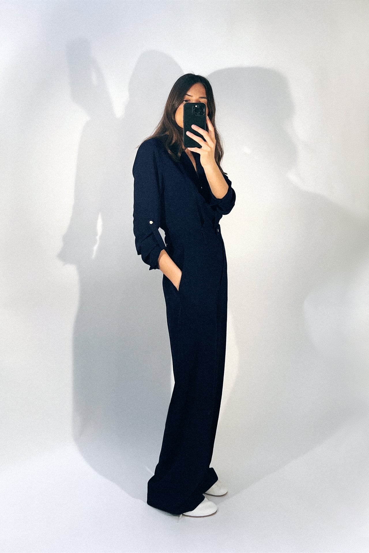 A person takes a mirror selfie, partially obscured by their phone, wearing a Zara wide-leg jumpsuit adorned with golden buttons. The navy color of the jumpsuit adds depth to the simplicity of the outfit, matched with understated white sneakers for a look that's both casual and chic.