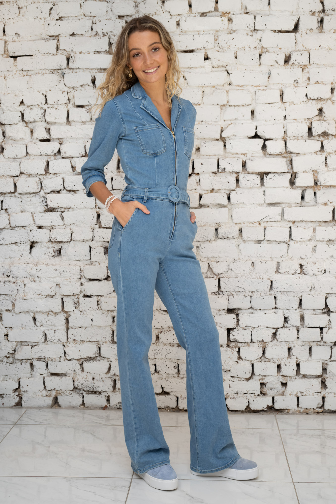 A radiant woman stands confidently against a textured white brick wall, wearing the White Coco Brooke Jumpsuit in a classic denim wash. The jumpsuit's flattering fit is accentuated by its cinched waist and flared legs, paired with casual white sneakers, embodying a relaxed yet chic aesthetic.