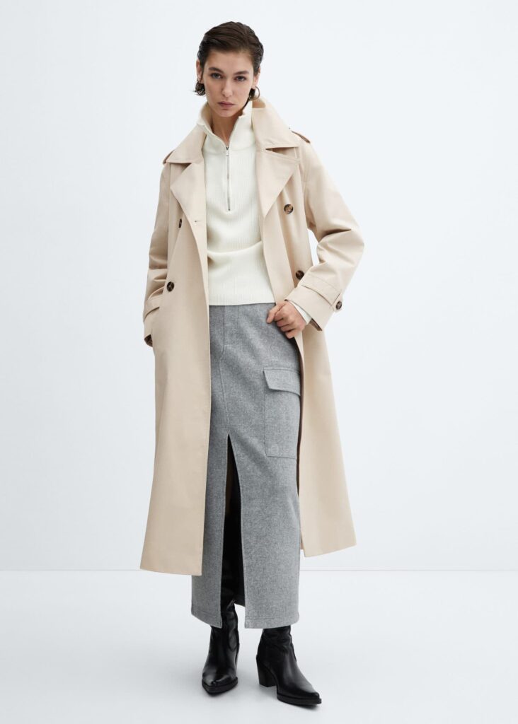 A model stands confidently in a Mango Double-button trench coat in Light Beige, layered over a snug white turtleneck and contrasting grey trousers. The ensemble is finished with black leather ankle boots, presenting a seamless blend of classic style and modern urbanity, ideal for a sophisticated yet practical city look.