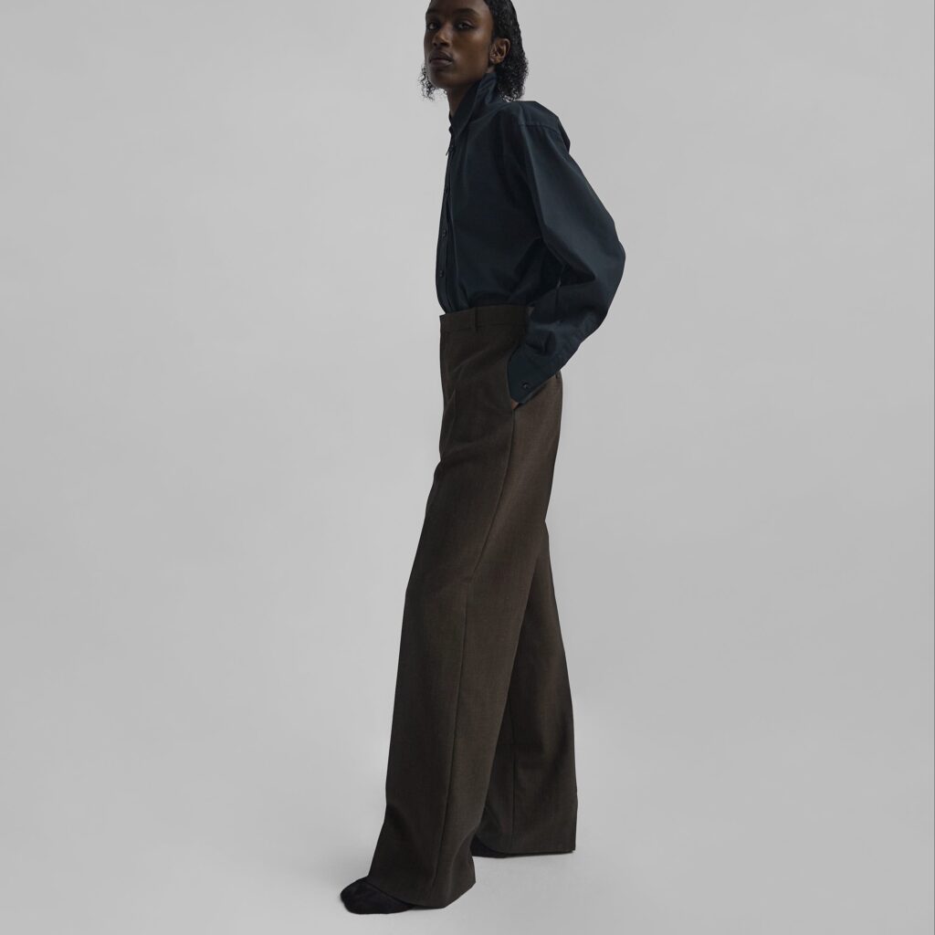A model poses in Phoebe Philo Milanese Trousers in a deep chocolate brown, paired with a dark, buttoned-up shirt for a look that's the epitome of understated chic. The trousers' wide-leg cut drapes elegantly, suggesting movement and a modern, tailored silhouette.
