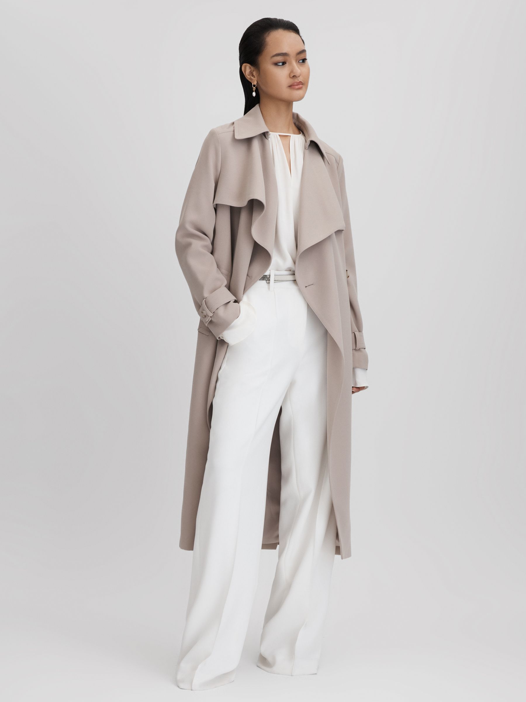 A poised model displays the Reiss Etta Double Breasted Trench in Mink Neutral, a garment that exudes effortless sophistication. The coat's soft hue complements the crisp white outfit underneath, creating a serene and polished look. The trench's fluid drape and the model's elegant stance suggest both comfort and timeless style.