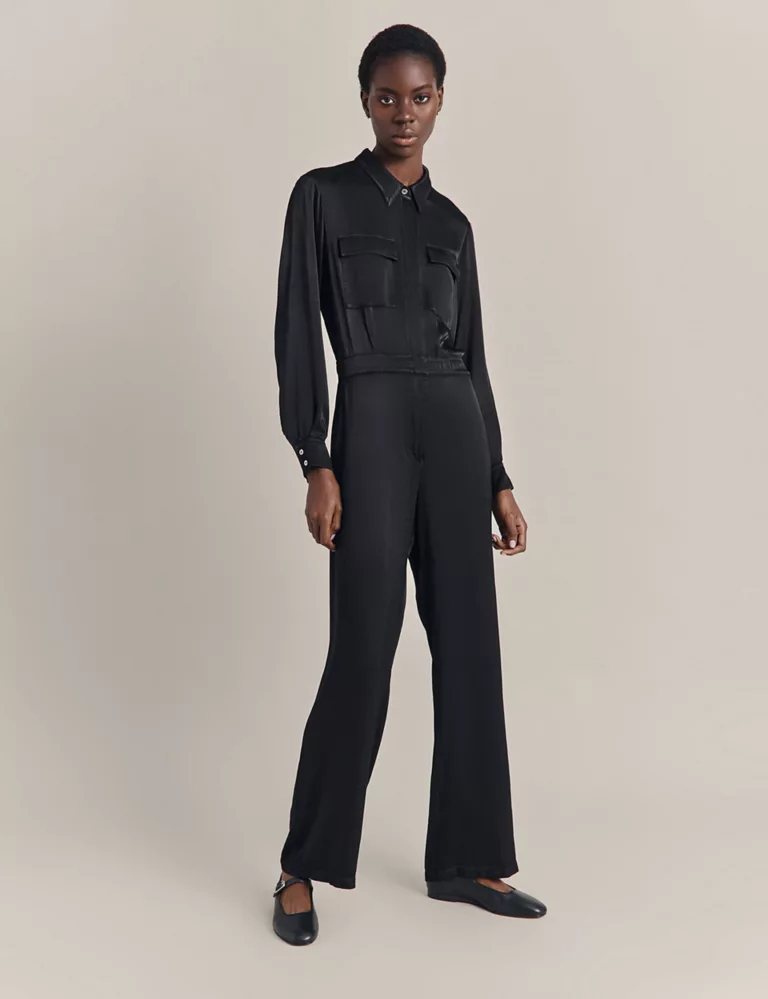 A model poses with an air of understated elegance in a Ghost Utility Pocket Long Sleeve Waisted Jumpsuit in Black. The jumpsuit's sleek design features practical pockets and a fitted waist, paired with classic black flats, encapsulating a modern take on utilitarian chic.