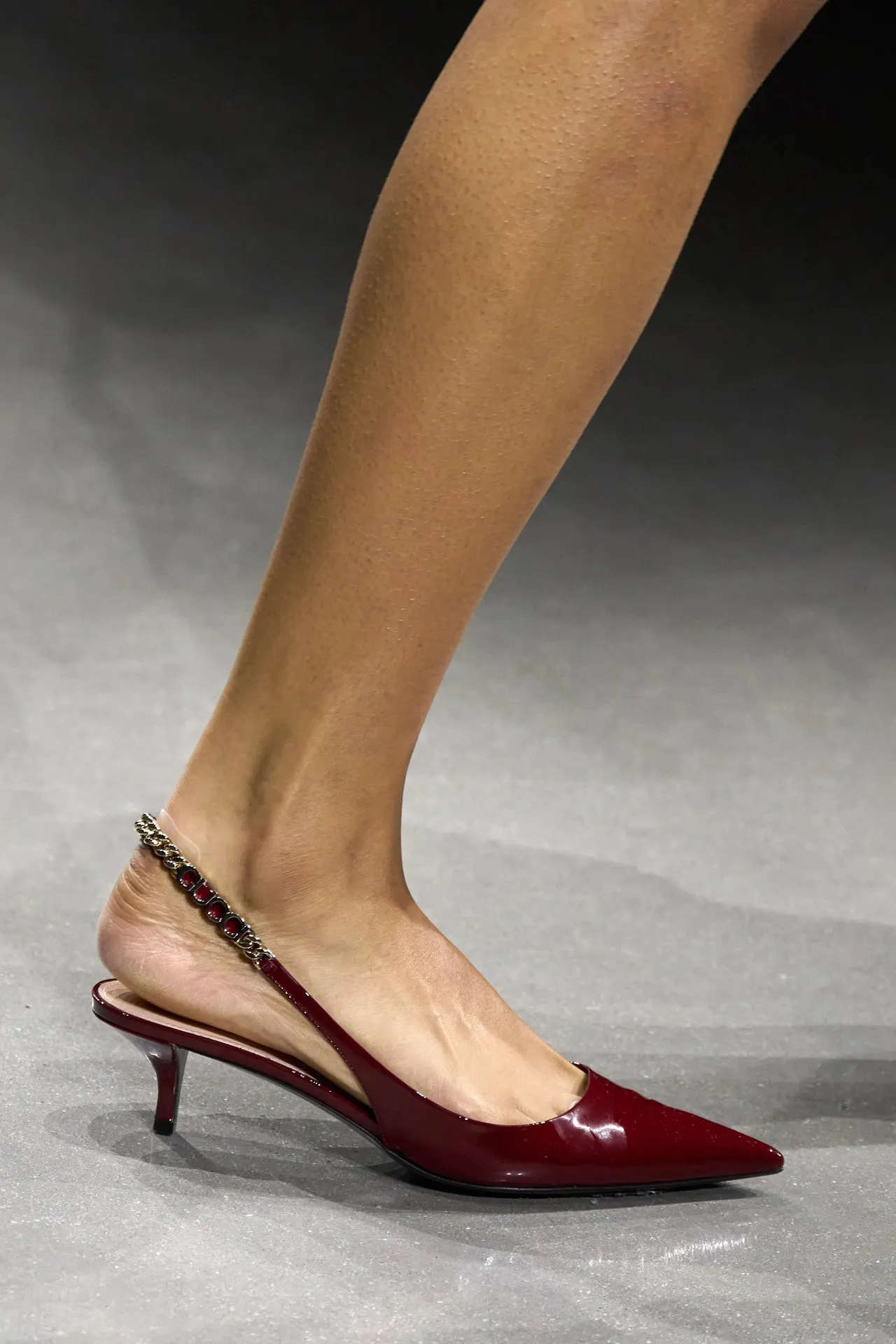An image capturing the elegance of a slender leg adorned with a glossy, oxblood-colored kitten heel. The shoe, with a pointed toe and delicate heel, features a charming embellishment - a sparkling anklet with crimson gems cradled in silver settings, adding a touch of glamour to every step. This snapshot is all about the grace of subtle details and the power of a shoe to not just complement an outfit but to define a moment.