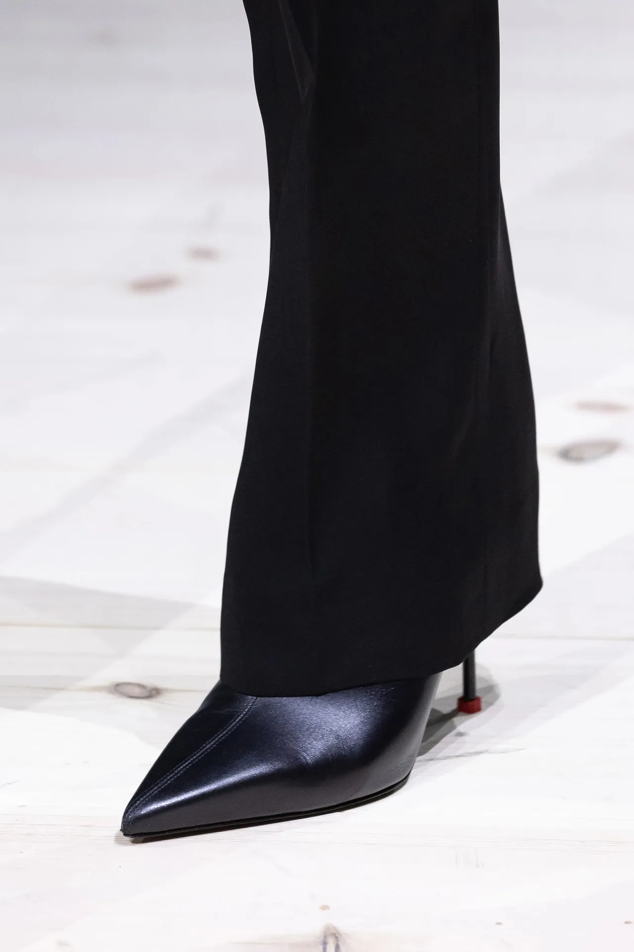 A snapshot of poise and sophistication: the image features a sleek, black pointed-toe shoe by Alexander McQueen peeking out from beneath the hem of tailored trousers. The shoe's silhouette is the epitome of elegance, its smooth leather surface catching the light just so. The subtle hint of a red sole adds an unexpected pop of colour, a signature nod to the world of high fashion. This is more than footwear; it's a statement, a piece of art that stands confidently on the wooden floor.