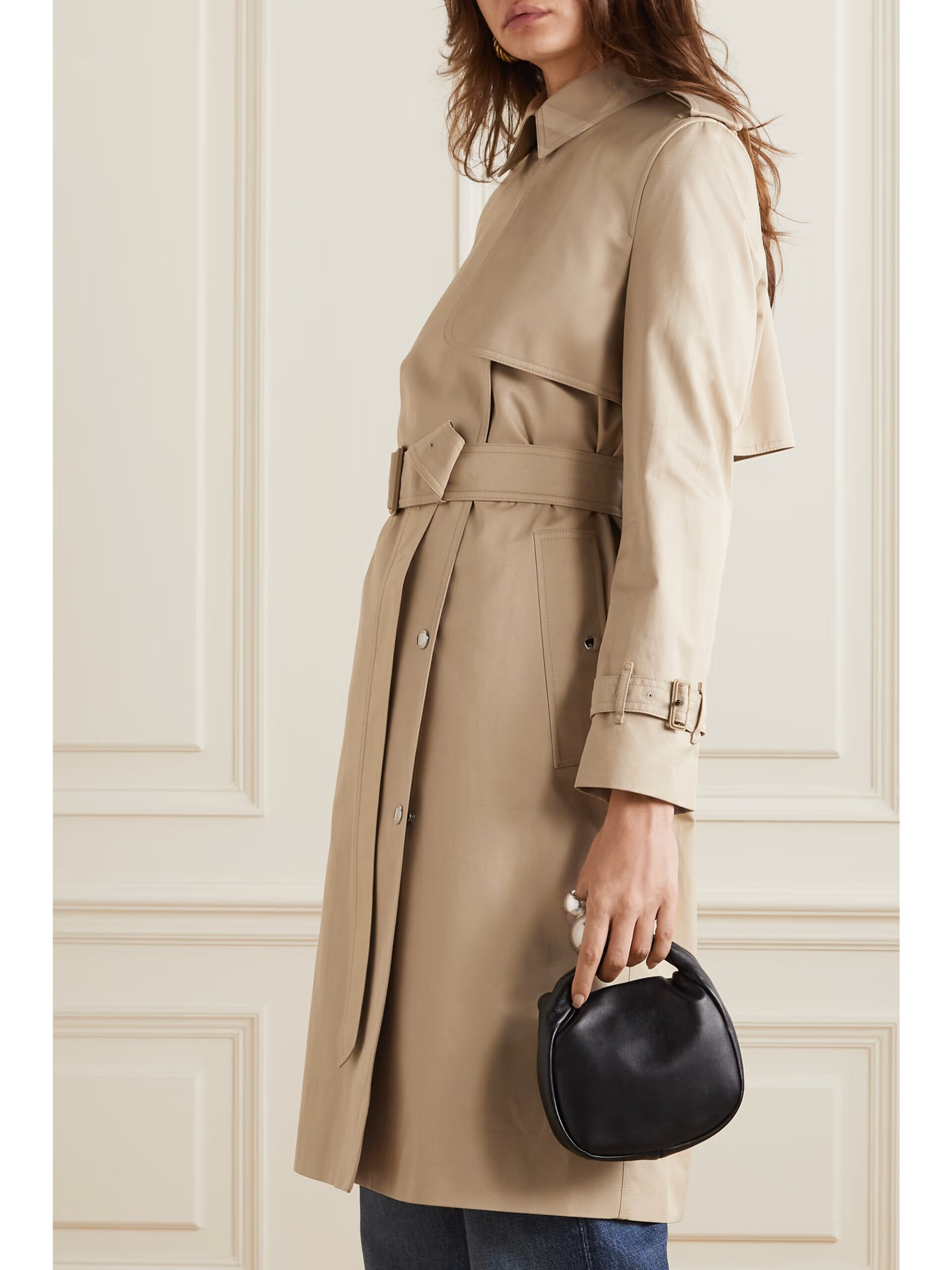 A woman stands side-on showcasing a BURBERRY Sandridge belted cotton-gabardine trench coat in a neutral shade. The coat's tailored silhouette is accentuated by a cinched waist belt and structured shoulders, complemented by a dark handheld bag, hinting at a blend of classic style with modern elegance.