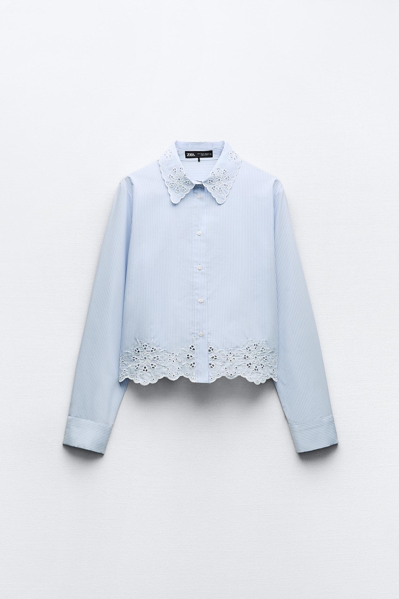 A cropped striped shirt from Zara, featuring light blue vertical stripes with a contrasting white, scalloped lace collar and hem. The shirt has a front button closure and full-length sleeves with buttoned cuffs. The piece is displayed on a neutral background, accentuating its delicate and feminine design.